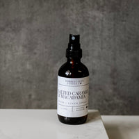 Bottle of 'Salted Caramel & Macadamia' room spray on a gray surface with a neutral background