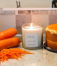 A carrot cake candle with a lit flame, placed on a countertop alongside whole carrots and a slice of carrot cake, with a cardboard label that reads 'Carrot Cake'.