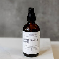 Bottle of 'Home Sweet Home' room and linen spray on a white surface with a gray background