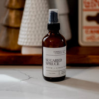Bottle of Sugared Spruce room and linen spray on a marble surface with decorative christmas items in the background.