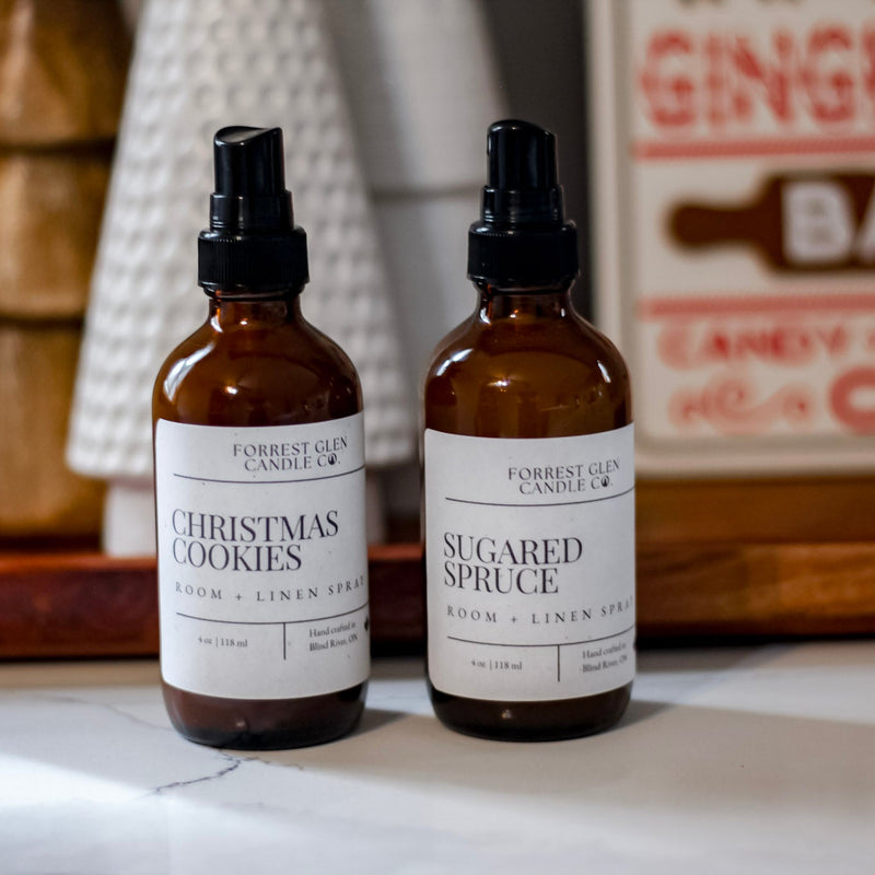 Two bottles of 'Christmas Cookies' and 'Sugared Spruce' room and linen sprays on a surface with decorative items in the background.