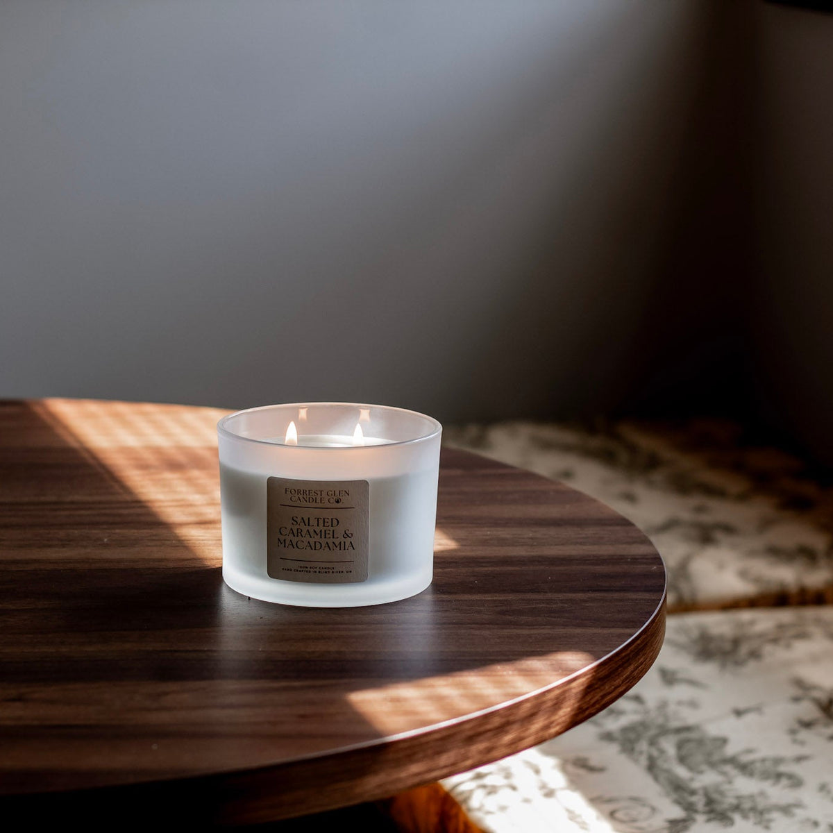 A frosted glass jar candle with a beige label sits on a dark wooden table in a softly lit interior setting.