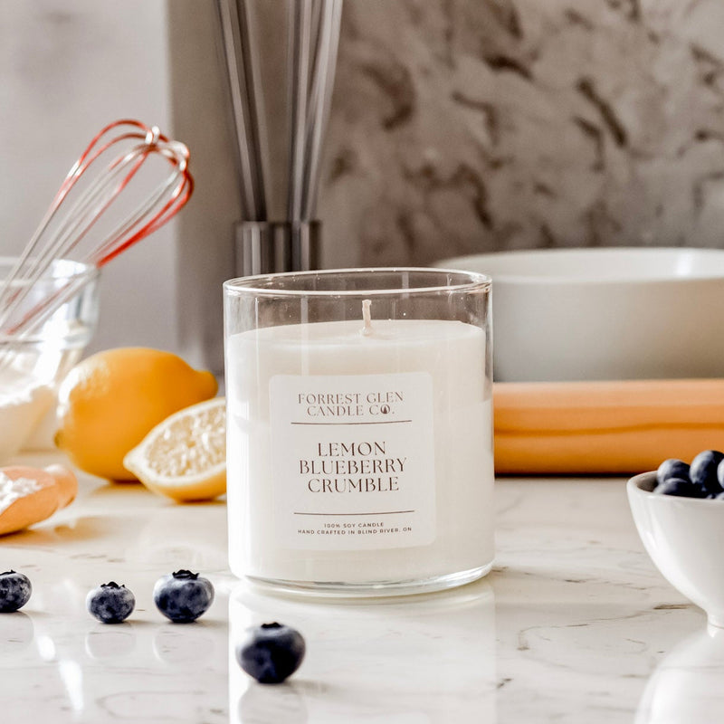 Candle labeled 'Lemon Blueberry Crumble' on a kitchen counter with lemons, blueberries, and baking utensils.