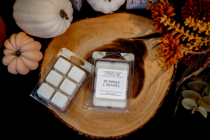 Two wax melts packages from Forrest Glen Candle Co. labeled 'Pumpkin Caramel,' placed on a round wood slice with decorative pumpkins and autumn flowers around them on a black background.