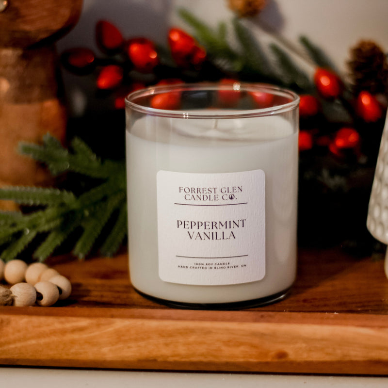 A soy wax candle with a label that reads 'Forrest Clinch Candle Co. - Peppermint Vanilla', placed on a wooden surface with a wreath and Christmas decorations in the background.