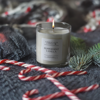 Candle labeled 'Peppermint Vanilla' with candy canes and greenery on a dark surface with blurred lights in the background