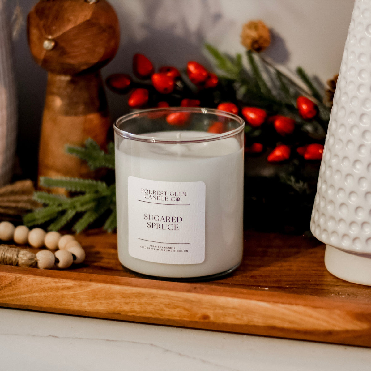 A soy wax candle with a label 'Forest Glen Candle Co.', placed on a wooden platter, with decorative items and a festive tree in the background.