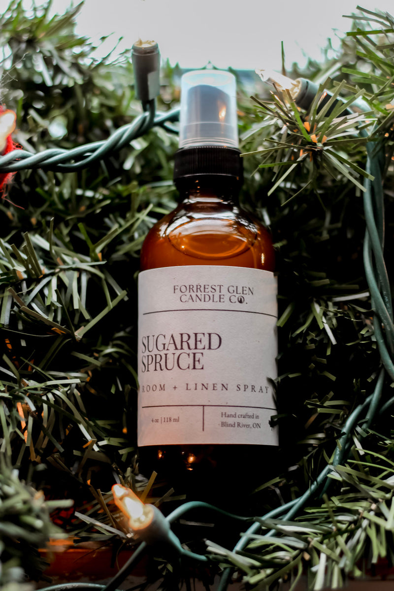 Bottle of Sugared Spruce room and linen spray from Forrest Glen Candle Co. on a green wreath with lights.