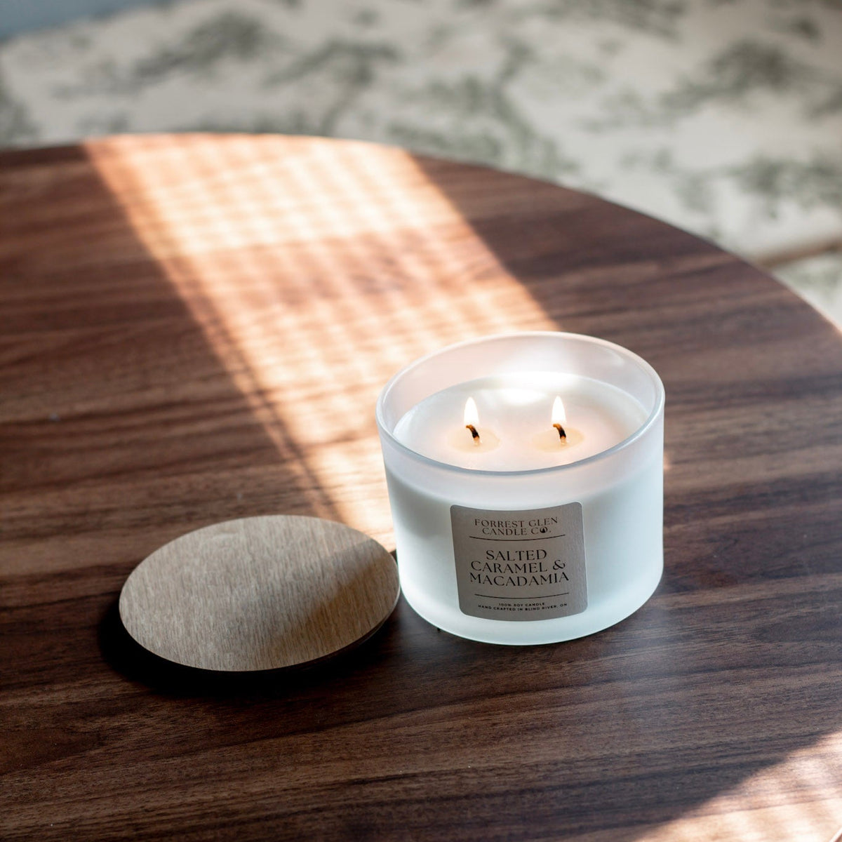 A lit white glass jar candle with two wicks sits on a dark wood table next to its round beige fabric lid. Sunlight casts a striped shadow across the tabletop, and a blurred patterned rug is visible in the background.