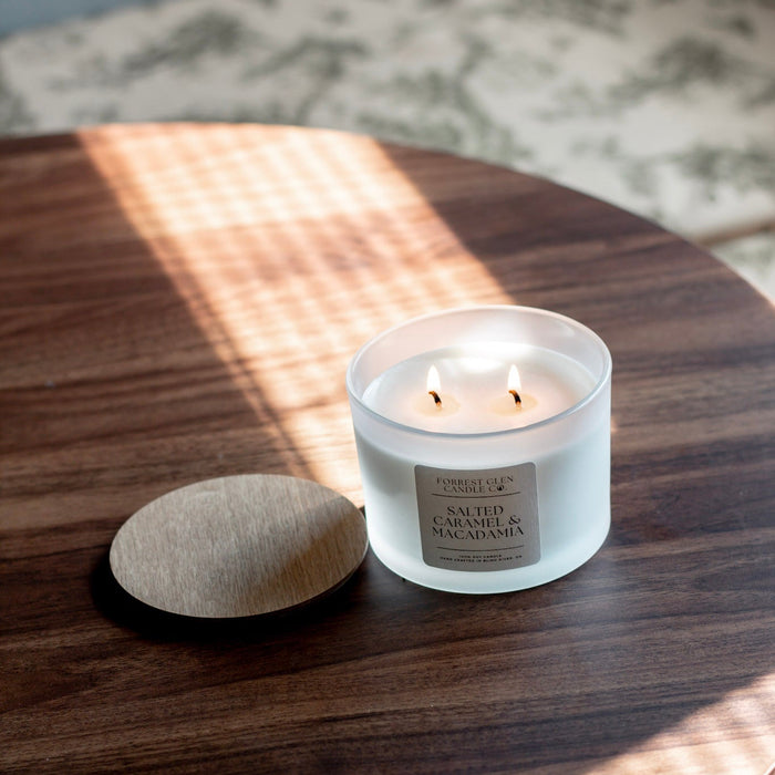 A lit white glass jar candle with two wicks sits on a dark wood table next to its round beige fabric lid. Sunlight casts a striped shadow across the tabletop, and a blurred patterned rug is visible in the background.